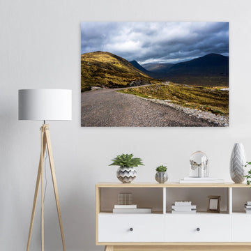 Glencoe Valley wall art showing a winding road from Glencoe Mountain Resort with golden hills, dark mountains and a dramatic cloudy sky over the Highland valley