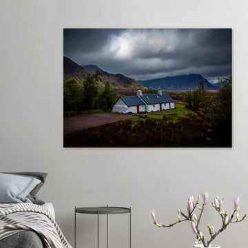 Blackrock Cottage Glencoe wall art showing a dark moody wide view of the white cottage surrounded by rough moorland and dramatic Scottish mountains under heavy clouds
