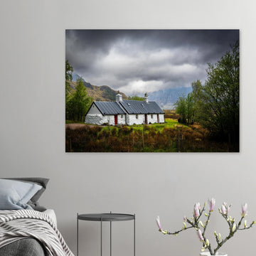 Blackrock Cottage Buachaille Etive Mòr wall art showing the white cottage in green moorland with trees and dramatic cloudy mountains in the Glencoe landscap