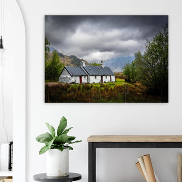 Blackrock Cottage Buachaille Etive Mòr wall art showing the white cottage in green moorland with trees and dramatic cloudy mountains in the Glencoe landscap