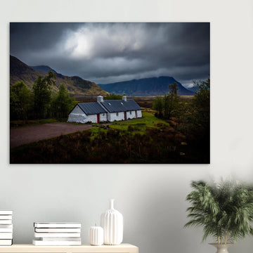 Blackrock Cottage Glencoe wall art showing a dark moody wide view of the white cottage surrounded by rough moorland and dramatic Scottish mountains under heavy clouds