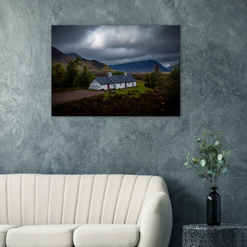 Blackrock Cottage Glencoe wall art showing a dark moody wide view of the white cottage surrounded by rough moorland and dramatic Scottish mountains under heavy clouds