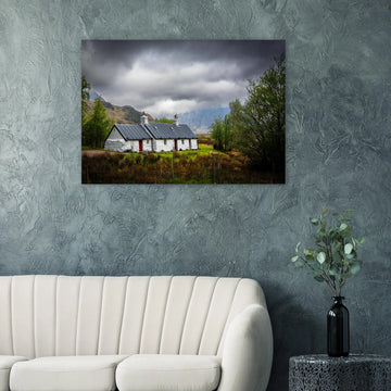 Blackrock Cottage Buachaille Etive Mòr wall art showing the white cottage in green moorland with trees and dramatic cloudy mountains in the Glencoe landscap
