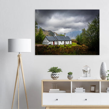 Blackrock Cottage Buachaille Etive Mòr wall art showing the white cottage in green moorland with trees and dramatic cloudy mountains in the Glencoe landscap