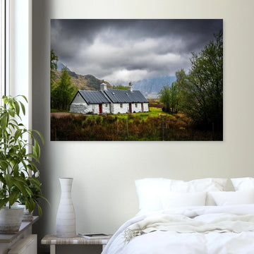 Blackrock Cottage Buachaille Etive Mòr wall art showing the white cottage in green moorland with trees and dramatic cloudy mountains in the Glencoe landscap