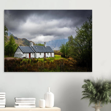 Blackrock Cottage Buachaille Etive Mòr wall art showing the white cottage in green moorland with trees and dramatic cloudy mountains in the Glencoe landscap