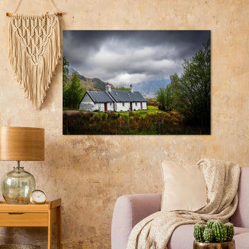 Blackrock Cottage Buachaille Etive Mòr wall art showing the white cottage in green moorland with trees and dramatic cloudy mountains in the Glencoe landscap