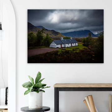 Blackrock Cottage Glencoe wall art showing a dark moody wide view of the white cottage surrounded by rough moorland and dramatic Scottish mountains under heavy clouds