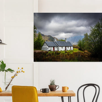 Blackrock Cottage Buachaille Etive Mòr wall art showing the white cottage in green moorland with trees and dramatic cloudy mountains in the Glencoe landscap