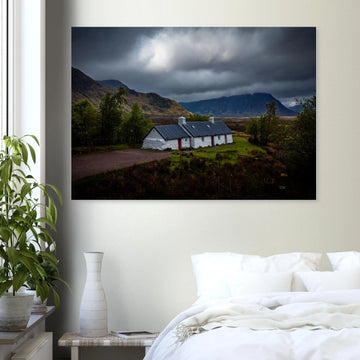 Blackrock Cottage Glencoe wall art showing a dark moody wide view of the white cottage surrounded by rough moorland and dramatic Scottish mountains under heavy clouds
