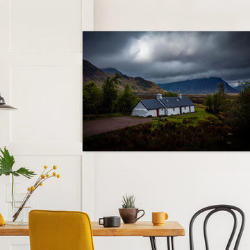 Blackrock Cottage Glencoe wall art showing a dark moody wide view of the white cottage surrounded by rough moorland and dramatic Scottish mountains under heavy clouds
